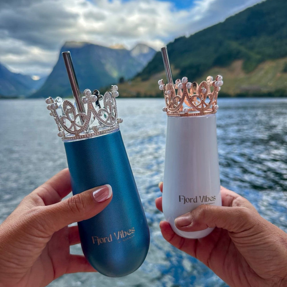 bilde av to hender som holder hver sin prosecco-kopp i hhv hvit og blå med her sin pyntekrone og sugerør. De skåler foran fjord og fjell