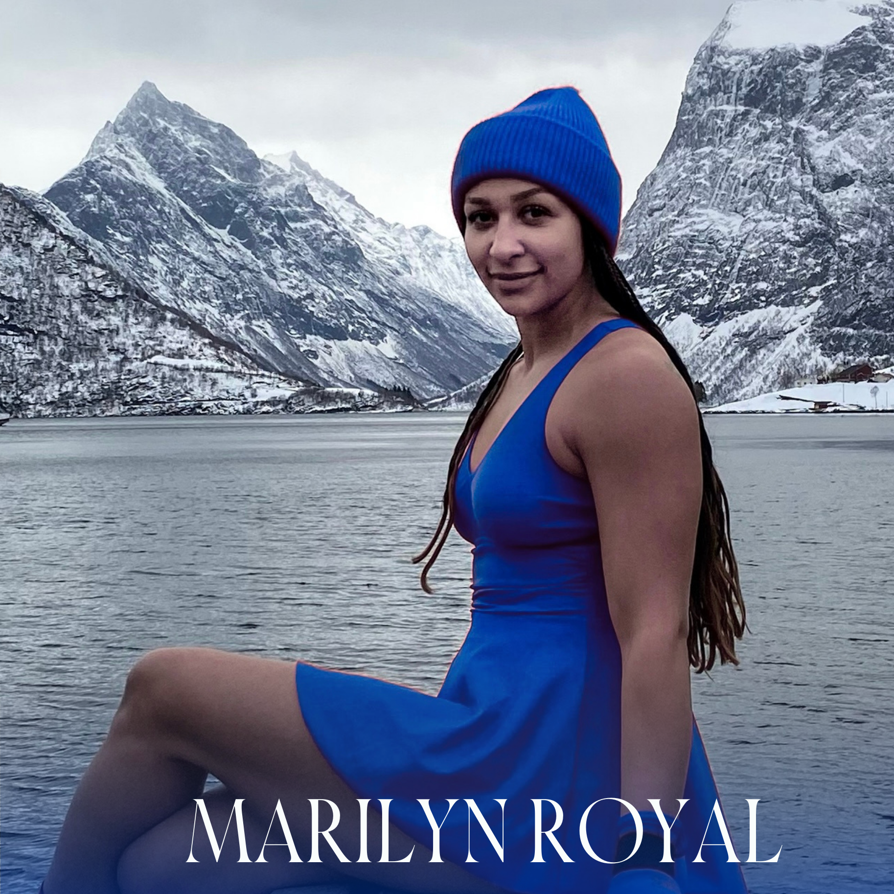 Marilyn - Fjord Vibes badekjole Kongeblå💙LOW CUT på en dame som sitter ved en fjord i norge med snøkledd e fjell. Hun har på matchende lue i ull