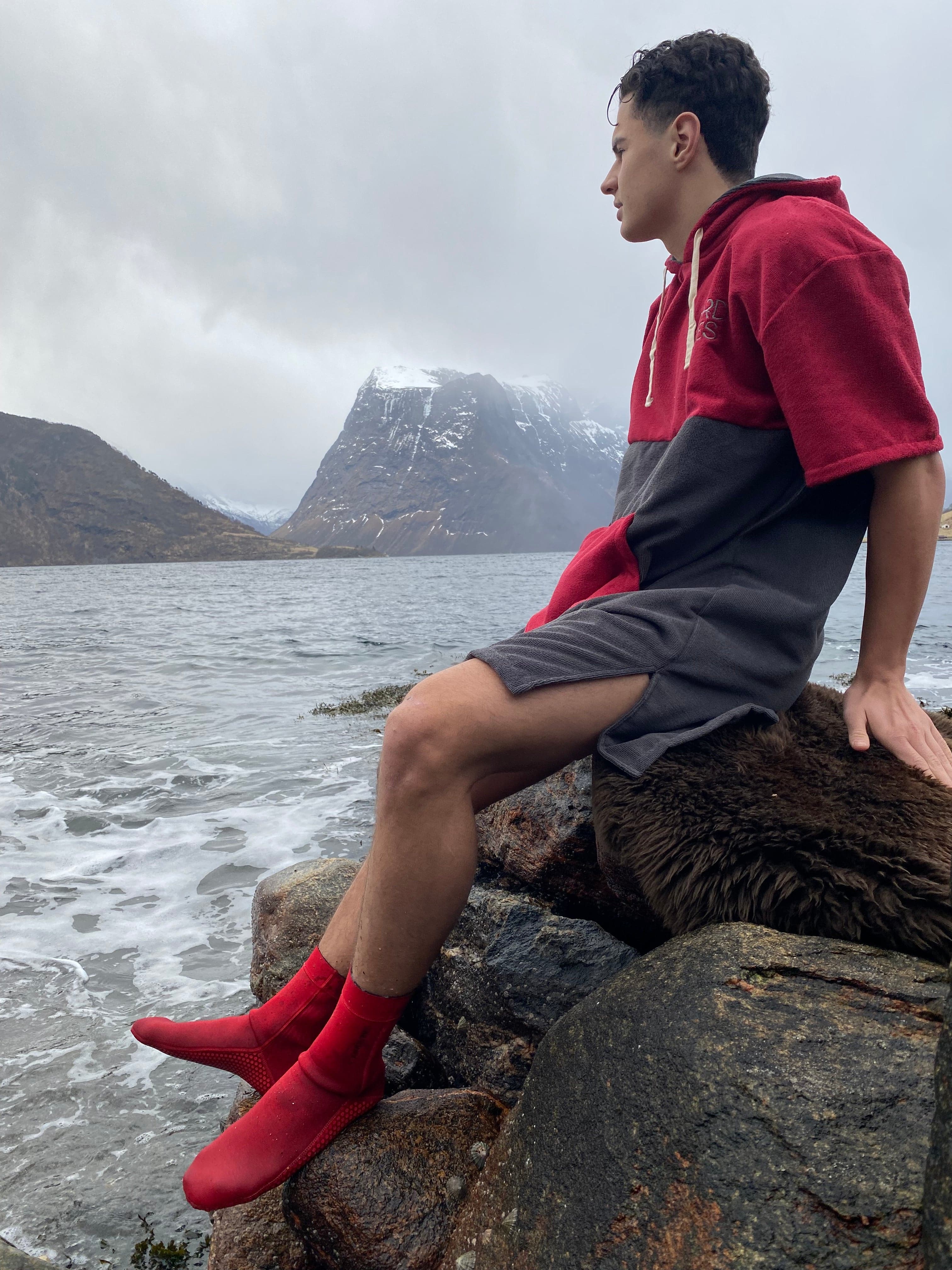 en mann som sitter i fjøresteinene med rød badeponcho og røde neoprensokker.