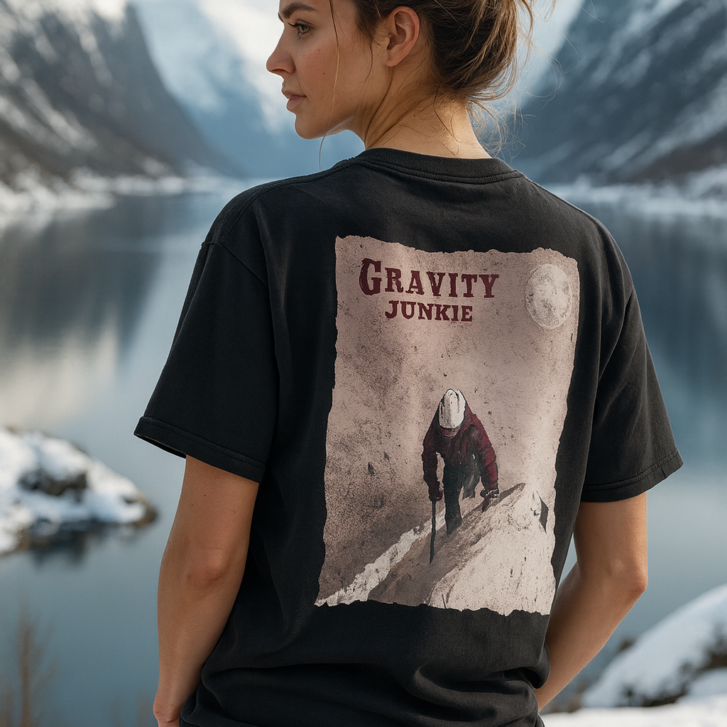 Ei dame som har på en randonnee t-skjorte i en oversized unies washed modell. Mørk grå med stort trykk på ryggen av en person som er på vei opp et fjell ved bruk av stegjern og is-øks